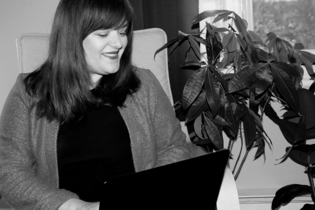 Une femme souriante assise confortablement dans son bureau, tenant un ordinateur portable. Elle est entourée par une ambiance chaleureuse et une plante verte luxuriante à sa droite. La photo est en noir et blanc, ce qui ajoute une touche classique et intemporelle à la scène